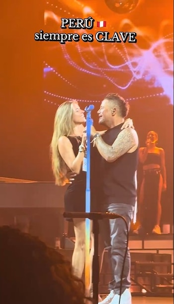 Alejandro Sanz y Stephanie Cayo se abrazan y cantan en un escenario iluminado con luces naranjas, con un micrófono azul entre ellos
