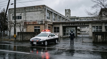 Un patrullero blanco y negro con luces rojas encendidas y un policía de espaldas frente al edificio de una escuela en un día nublado.