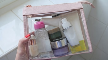 Una mano sostiene un neceser transparente con borde rosa que contiene frascos, tubos y botes de cosméticos sin etiquetas visibles.