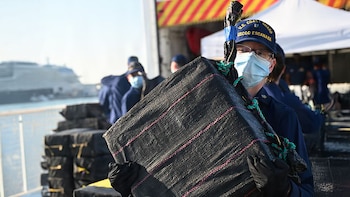Primer plano de un oficial de la Guardia Costera de EE. UU. con gorra y mascarilla, llevando un fardo grande negro. Al fondo, más fardos y personal en un muelle con un barco