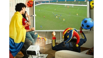 Tres personas observan un partido de fútbol en un proyector, con bandera de Colombia, vuvuzela, sombrero de fútbol y balones inflables
