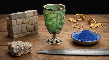 Vista aérea de objetos históricos sobre una mesa de madera: una copa verde, una estructura de bloques, joyas de oro, un cuenco con polvo azul, un trozo de hormigón y una hoja de acero.