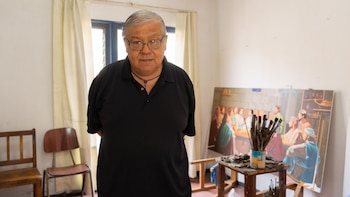 Retrato de un hombre de cabello gris y gafas con polo negro y collar púrpura, de pie en un estudio con dos sillas, un caballete, un cuadro y pinceles