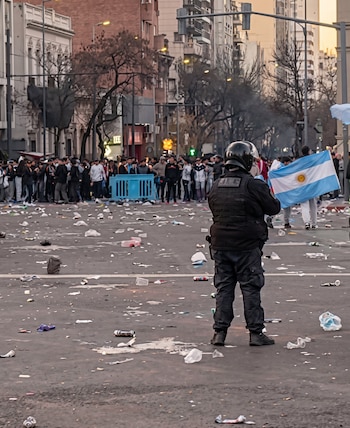 Oficial de policía de espalda en calle con basura. Al fondo, multitud tras valla azul y personas con bandera de Argentina. Edificios a los lados