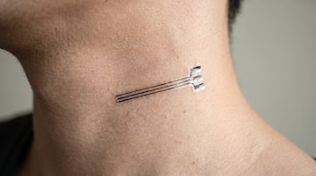 Primer plano del cuello de una persona con vello fino. Un pequeño sensor biométrico transparente con tres líneas conductoras oscuras se adhiere a la piel.