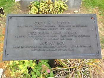 Placa de metal oscura con texto grabado que detalla datos de la familia Bates, rodeada de césped y algunas plantas verdes y secas