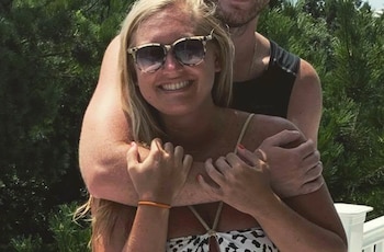 Mujer rubia de pelo largo con gafas de sol sonríe mientras es abrazada por detrás por un hombre. Se ven árboles verdes y una barandilla blanca de fondo
