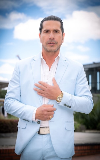 Gregorio Pernía viste un traje de color azul claro, camisa blanca y reloj dorado en un espacio exterior con cielo nublado
