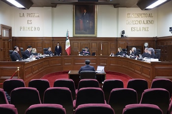 Una sala de audiencias con jueces de la Suprema Corte sentados en un estrado curvo de madera. Banderas, un retrato y asientos vacíos en primer plano