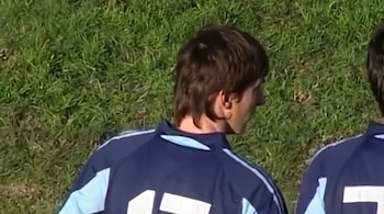 Un joven futbolista de cabello castaño viste una prenda deportiva azul sobre un campo de césped.