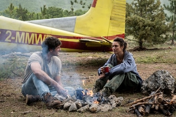Un hombre y una mujer sentados frente a una fogata encendida con una cafetera. Detrás hay un avión amarillo y rojo estacionado y árboles