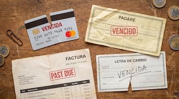 Vista aérea de una tarjeta de crédito rota, un pagaré, una factura con sello "PAST DUE" y una letra de cambio con "VENCIDA" en una mesa de madera.