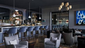 Una barra de bar con taburetes altos tapizados en azul frente a una zona de estar con sillones grises y lámparas de techo