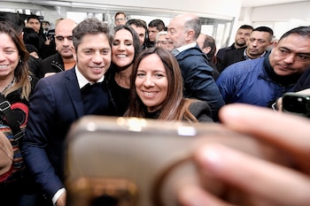 Axel Kicillof sonriente entre una multitud, con un teléfono móvil en primer plano que es sostenido por una mano en la parte inferior