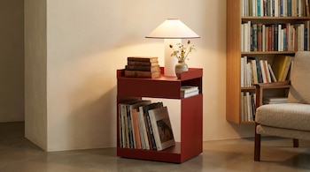Una mesa auxiliar de metal rojo con dos niveles, una lámpara, una pila de libros y un jarrón con flores, junto a una estantería de madera y un sillón beige.