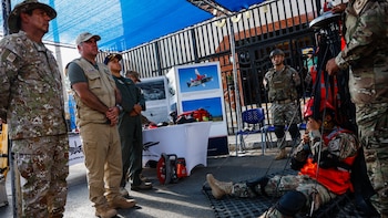 Hombres con uniformes militares y de emergencia observan a un hombre en una camilla de rescate con arneses, junto a paneles de aviones y una sierra