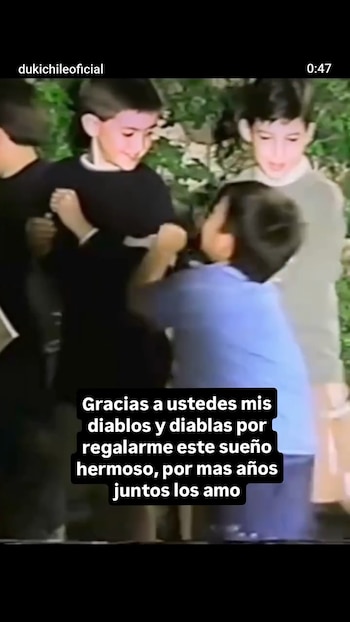 Cuatro niños pequeños juegan al aire libre, uno viste camiseta negra, otro camiseta azul. Hay vegetación de fondo y texto sobre la imagen