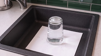 Vaso de vidrio transparente invertido sobre una hoja de papel blanco húmeda dentro de un fregadero de cocina oscuro con grifo de metal