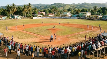 Vista aérea de un partido de béisbol con jugadores en un diamante de tierra y césped. Una multitud de espectadores rodea el campo bajo un cielo claro y colinas verdes.