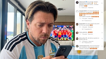 Un hombre con barba mira un teléfono móvil mientras lleva una camiseta de fútbol de Argentina. Una pantalla de televisión con un partido y una bandera argentina se ven al fondo.