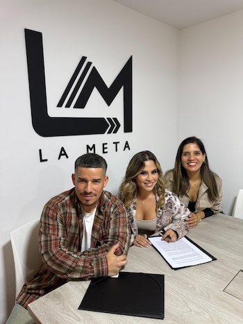 Tres personas, dos mujeres y un hombre, sentadas en una mesa frente a un logo negro con las letras LM y el texto LA META en una pared blanca
