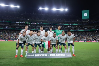 Once futbolistas de la selección mexicana posan en dos filas en la cancha de un estadio iluminado frente a un letrero de México contra Colombia