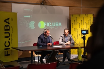 Dos hombres sentados frente a micrófonos en una mesa, con pendones amarillos y una pantalla proyectada con la marca FIC.UBA
