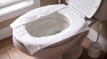 Un inodoro blanco con una cubierta de papel en el asiento, manchas de cal y sarro en el interior de la taza, y un cepillo de baño en su soporte en el suelo.