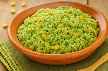 Recipiente de barro con arroz verde y granos de maíz amarillo sobre una servilleta verde y una mesa de madera.