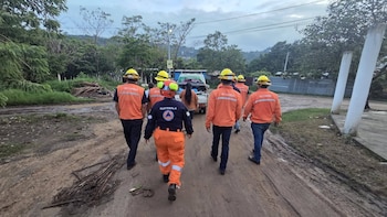 Un grupo de seis personas con uniformes naranjas y cascos amarillos camina por un camino de tierra embarrado. Una camioneta blanca y vegetación se ven al fondo