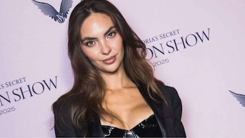 Natalie Vertiz, mujer de cabello castaño y largo, sonríe levemente con un top bustier negro y chaqueta, frente a un fondo rosa con el logo de Victoria's Secret
