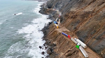 Vista aérea de una hilera de camiones detenidos en un camino costero sin pavimentar, ubicado entre el mar y una ladera de tierra