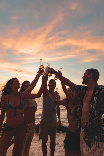 Varios jóvenes con ropa de playa levantan botellas de cerveza frente al mar durante el atardecer