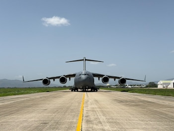 Avión militar de carga Boeing C-17 Globemaster III color gris en una pista de aterrizaje con línea amarilla central. Se ven pasto, montañas y edificios de hangar