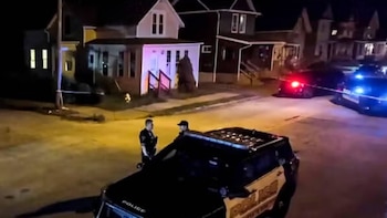Dos policías en uniforme conversan junto a un patrullero en una calle residencial nocturna con casas y luces de emergencia de fondo