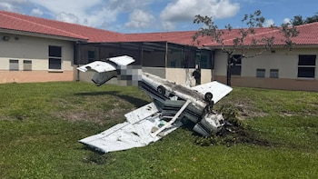 Avioneta blanca y azul volcada en un césped verde, con las alas rotas y la cola levantada, frente a un edificio con techo de tejas rojas y ventanas