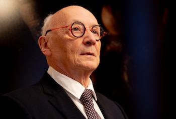 Primer plano de un hombre calvo y canoso de edad avanzada, con gafas de montura redonda roja y negra, vistiendo un traje oscuro y corbata estampada, con un pin negro en la solapa