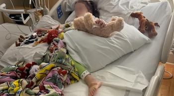Niño acostado en una cama de hospital con manta colorida, oso de peluche, otros juguetes, equipo médico, vendaje en el pie y pulsera identificativa