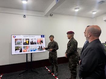 Dos militares en uniforme camuflado escuchan a un técnico con guantes de trabajo frente a estantes metálicos con pistolas en estuches