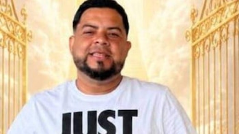 Hombre con barba y camiseta blanca con la palabra "JUST", sonriendo. Detrás, una puerta dorada ornamental