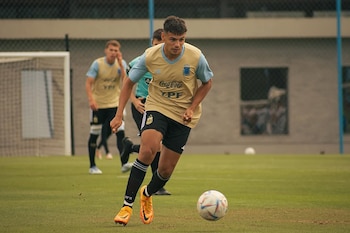Un futbolista de la selección argentina con camiseta beige y shorts negros corre en un campo de césped verde con un balón