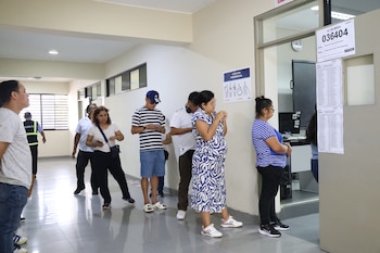 Varias personas hacen fila en un pasillo interior. Una mujer embarazada, un hombre con gorra y otros ciudadanos esperan. Hay un cartel de "Atención Preferencial"