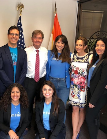 Siete personas, cinco de pie y dos arrodilladas, posan sonriendo frente a banderas y un sello oficial en una oficina