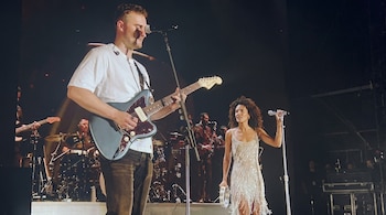 Un hombre toca una guitarra eléctrica frente a un micrófono, una mujer canta en un micrófono y sostiene una pandereta, músicos de fondo con batería y saxofón en un escenario.