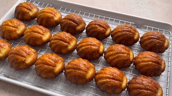 Dieciocho panecillos redondos de color dorado con superficie acanalada, colocados en una rejilla metálica sobre una bandeja blanca.