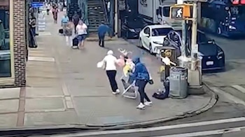 Personas caminando por una acera junto a un semáforo peatonal, un papelero y varios automóviles estacionados