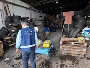 Múltiples bolsas de basura negras apiladas en el suelo de un pasillo, rodeadas por estanterías llenas de botellas de bebidas y un cartel de Aduana