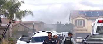 Varios agentes y vehículos policiales junto a una ambulancia frente a casas residenciales con una columna de humo en el fondo