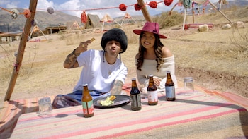 Wendy Sulca y Micro TDH sentados en una manta rayada con botellas de cerveza y comida al aire libre. Él con sombrero negro y ella con sombrero rojo. Fondo montañoso