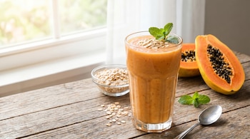 Vaso de batido de papaya con avena y menta. Mitades de papaya, cuenco de avena y cuchara sobre mesa de madera. Ventana luminosa de fondo.